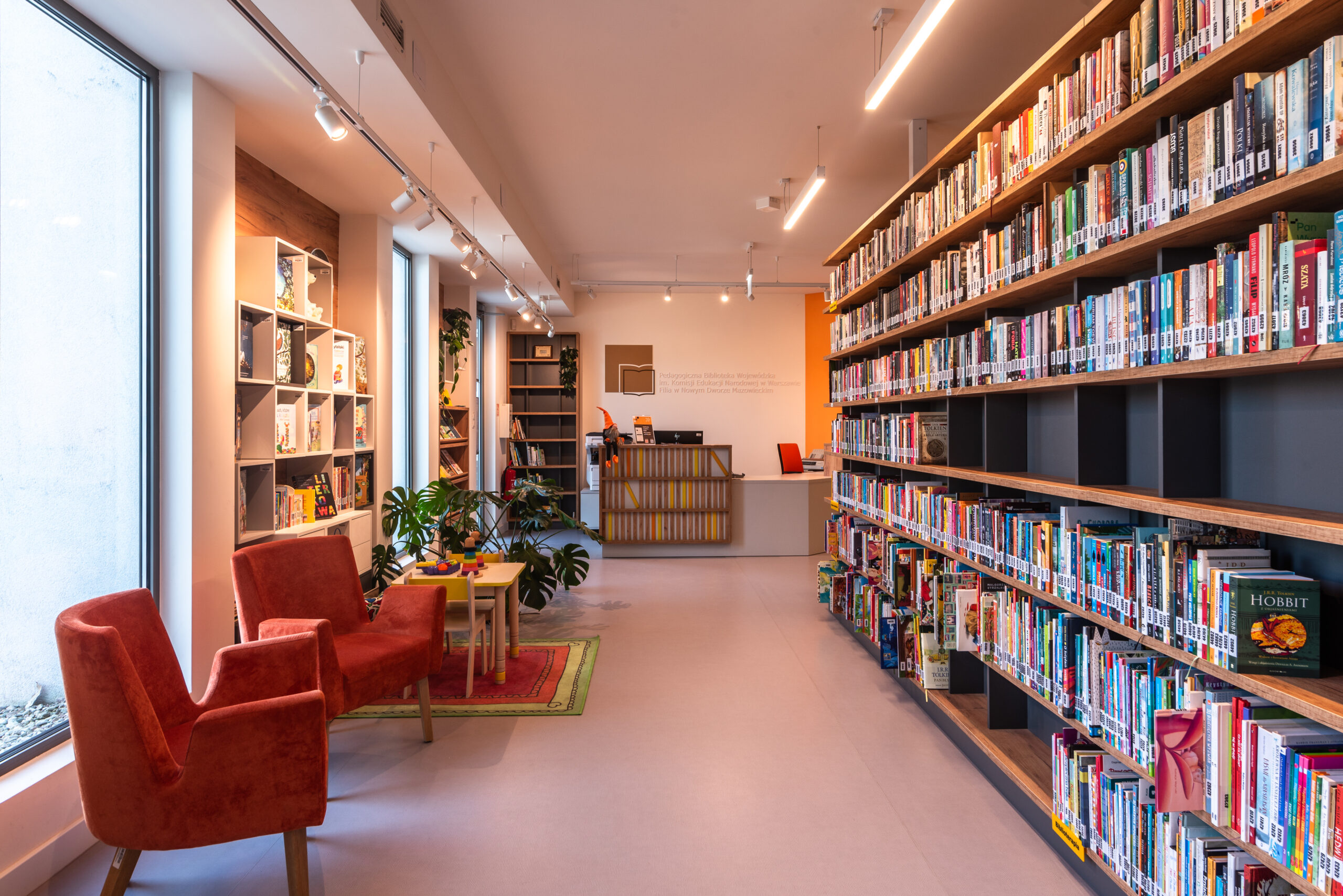 Nowoczesne, długie pomieszczenie biblioteki z dużymi oknami po lewej stronie, przez które wpada naturalne światło. Wzdłuż prawej ściany ciągnie się wysoki, ciemny regał z gęsto ułożonymi książkami. W głębi widoczna jest lada informacyjna na tle pomarańczowej ściany. W strefie wypoczynkowej przy oknie ustawiono dwa komfortowe, welurowe fotele w kolorze koralowym oraz mały stolik dziecięcy z krzesełkami